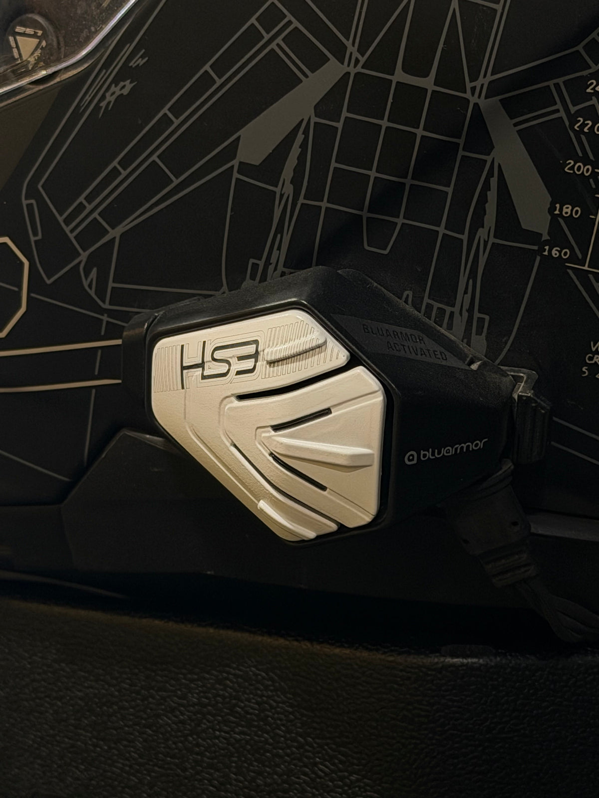 BluArmor HS3 helmet Bluetooth device with a silver faceplate against a dark patterned backdrop