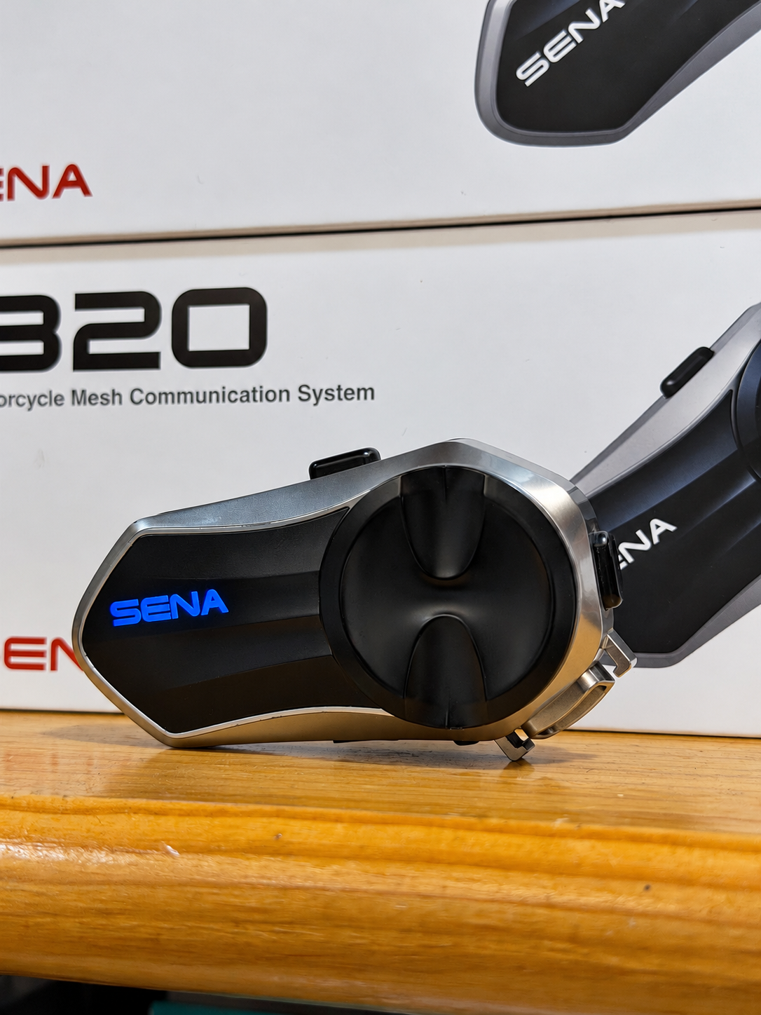 Sena B20 Mesh motorcycle intercom with illuminated blue Sena logo, displayed beside its packaging
